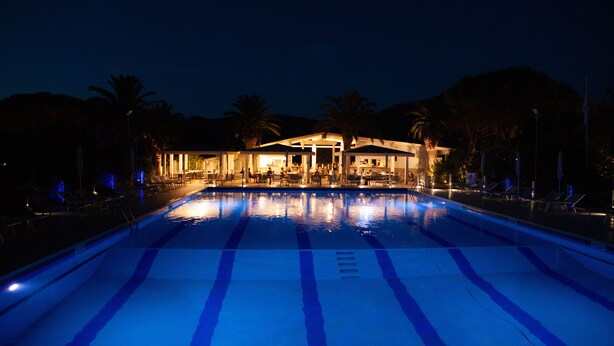 Una piscina con marcas azules que delimitan carriles, rodeada de sillones de descanso y palmeras, con un edificio al fondo.