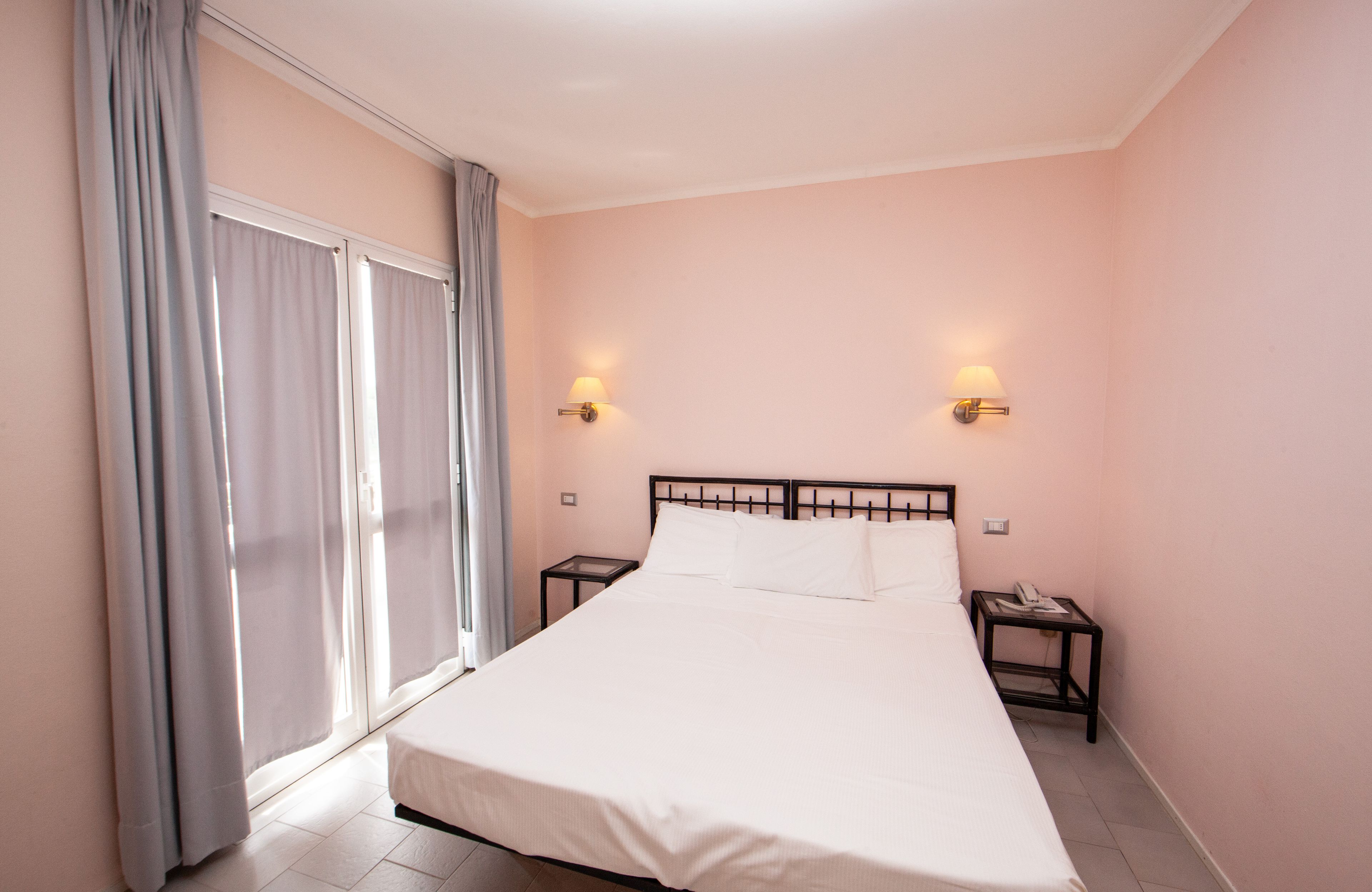A hotel room with a large bed, two bedside tables, a window with curtains, and wall-mounted lights.