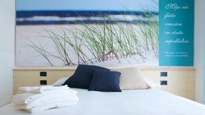 Un lit bien fait, avec du linge de lit blanc et deux oreillers, une œuvre d’art représentant une scène de plage accrochée au mur, et une enseigne de couleur turquoise avec du texte.