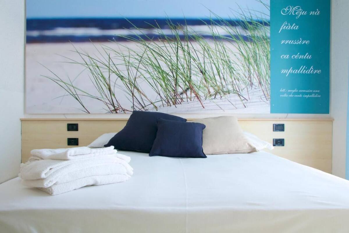 Ein ordentlich bezogenes Bett mit weißen Leinen und zwei Kissen, ein Wandbild mit einer Strandansicht sowie ein türkisblaues Schild mit Text.
