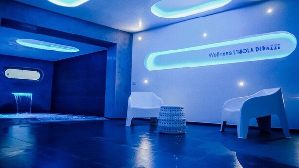 Un centre de bien-être moderne aux couleurs bleues, comprenant un espace d’accueil agrémenté d  ’un élément décoratif aquatique, deux chaises blanches et un tabouret à texture particulière.