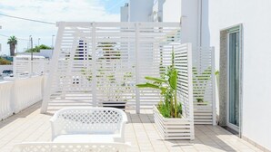 Un espace extérieur aménagé avec des sièges blancs, un pergolas blanc, un bac à fleurs contenant des plantes vertes et un mur blanc avec une fenêtre.