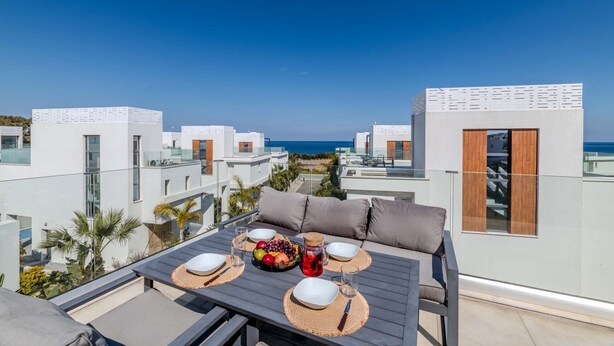 Une terrasse sur le toit aménagée avec un espace repas, comprenant une table avec un panier de fruits, des verres et une bouteille. La terrasse offre une vue sur une zone côtière avec des bâtiments modernes et un ciel bleu dégagé.
