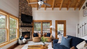 A cozy living room with a stone fireplace, wooden ceiling beams, and large windows offering a view of the outdoors.