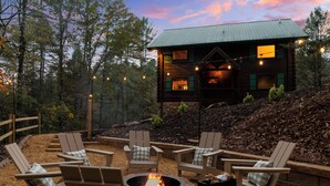 A cabin with a stone fire pit surrounded by Adirondack chairs, set in a forested area with a view of the cabin.