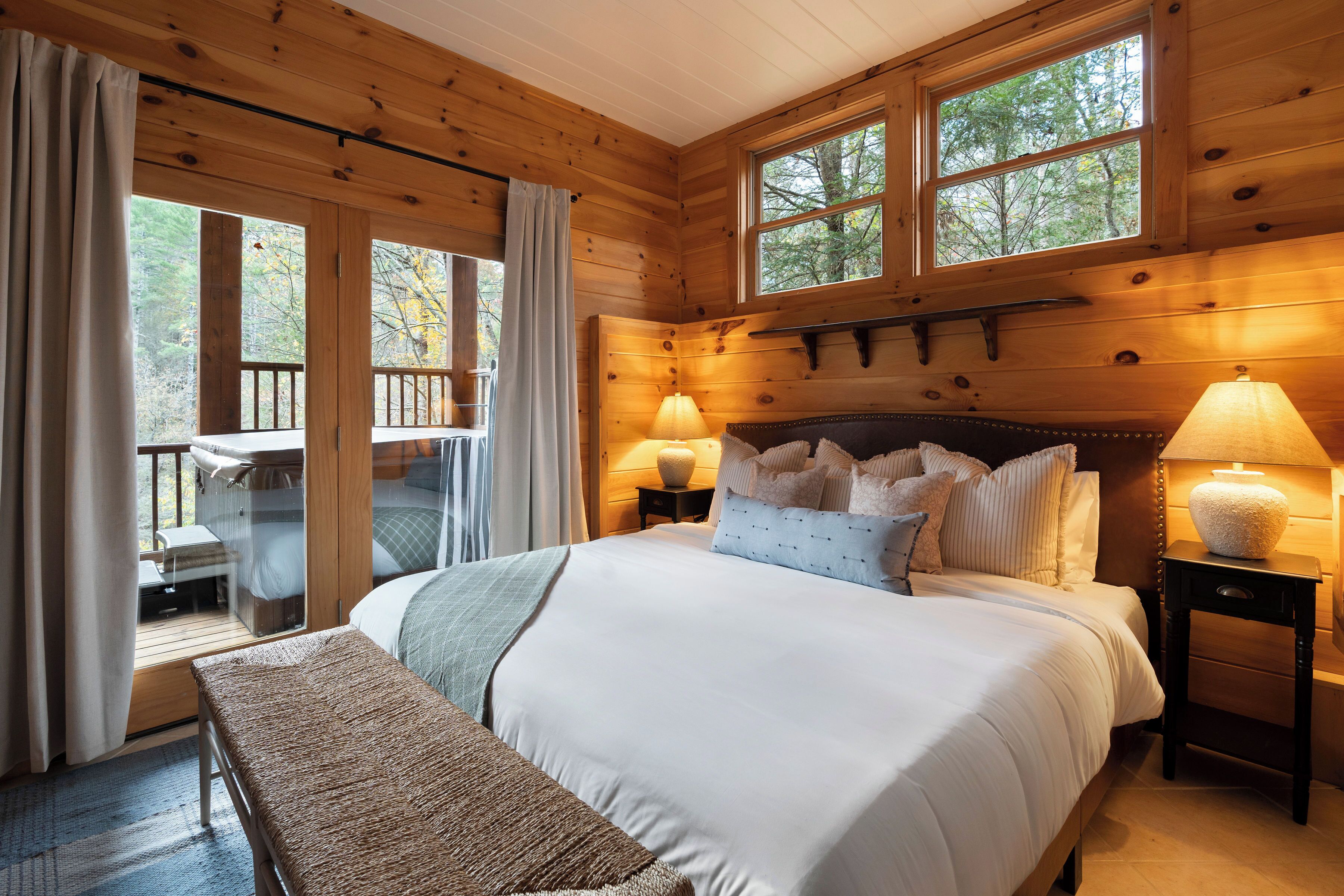Een slaapkamer in een houten blokhut met een groot bed, twee bedlampen, een bankje en uitzicht op de natuur door een schuifdeur.
