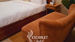 Una camera d'albergo con un letto in legno, biancheria bianca, un comodino con una sveglia e una poltrona marrone.