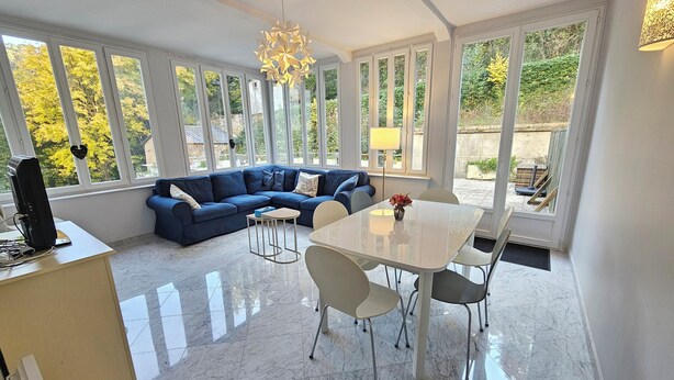 Ein modernes Wohnzimmer mit einem blauen Ecksofa, einem Esstisch mit Stühlen und großen Fenstern mit Blick auf Grünflächen.