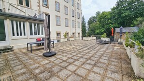 Ein Innenhof mit Fliesenboden, Gartenmöbeln und einem Gebäude mit mehreren Fenstern und Fensterläden.