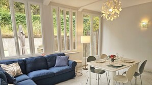 Ein modernes Wohnzimmer mit einem blauen Sofa, einem Esstisch für zwei Personen und großen Fenstern mit Blick auf Grünflächen.
