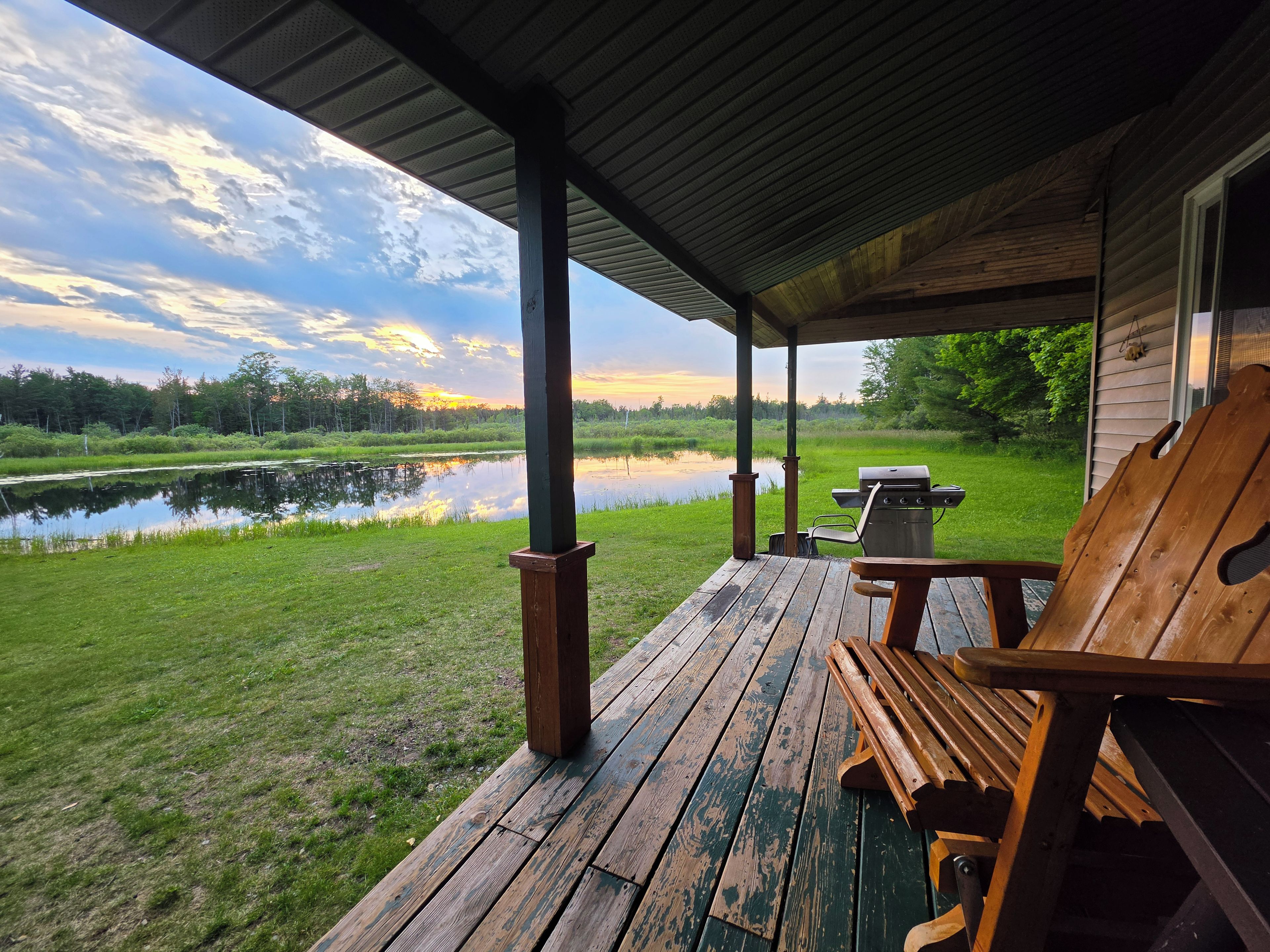Una terraza de madera con una parrilla y sillas, con vistas a un estanque y un bosque.