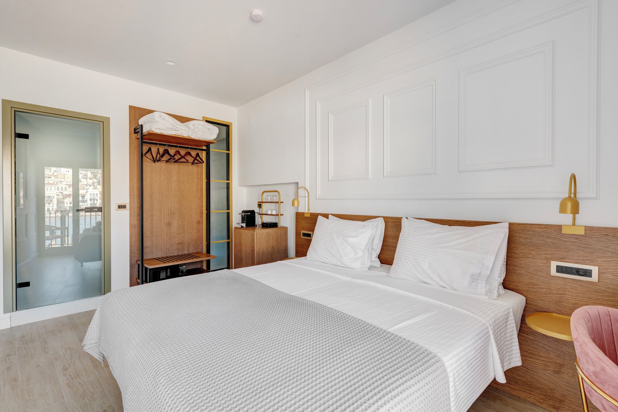 Ein modernes Hotelzimmer mit einem großen Bett, einem Holz-Kopfteil, einem Nachttisch mit Lampe und Blick auf die Stadt durch eine Glasdoor.