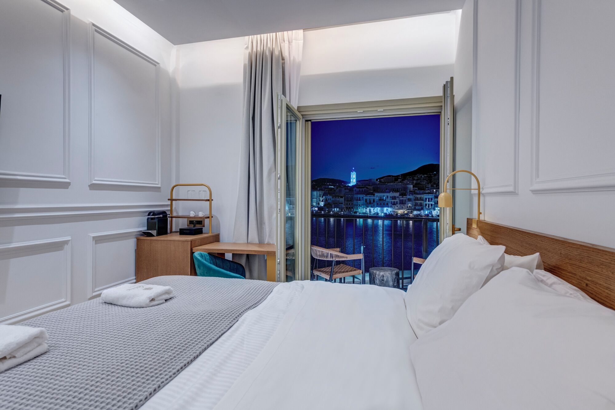 Ein Hotelzimmer mit Bett, Schreibtisch mit Lampe, Stuhl und Blick auf eine nächtliche Stadtlandschaft.