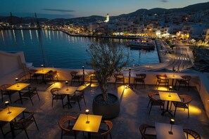 An outdoor dining area with tables and chairs, overlooking a body of water and a cityscape at dusk. - Eclectic Syros (Ermoupoli)