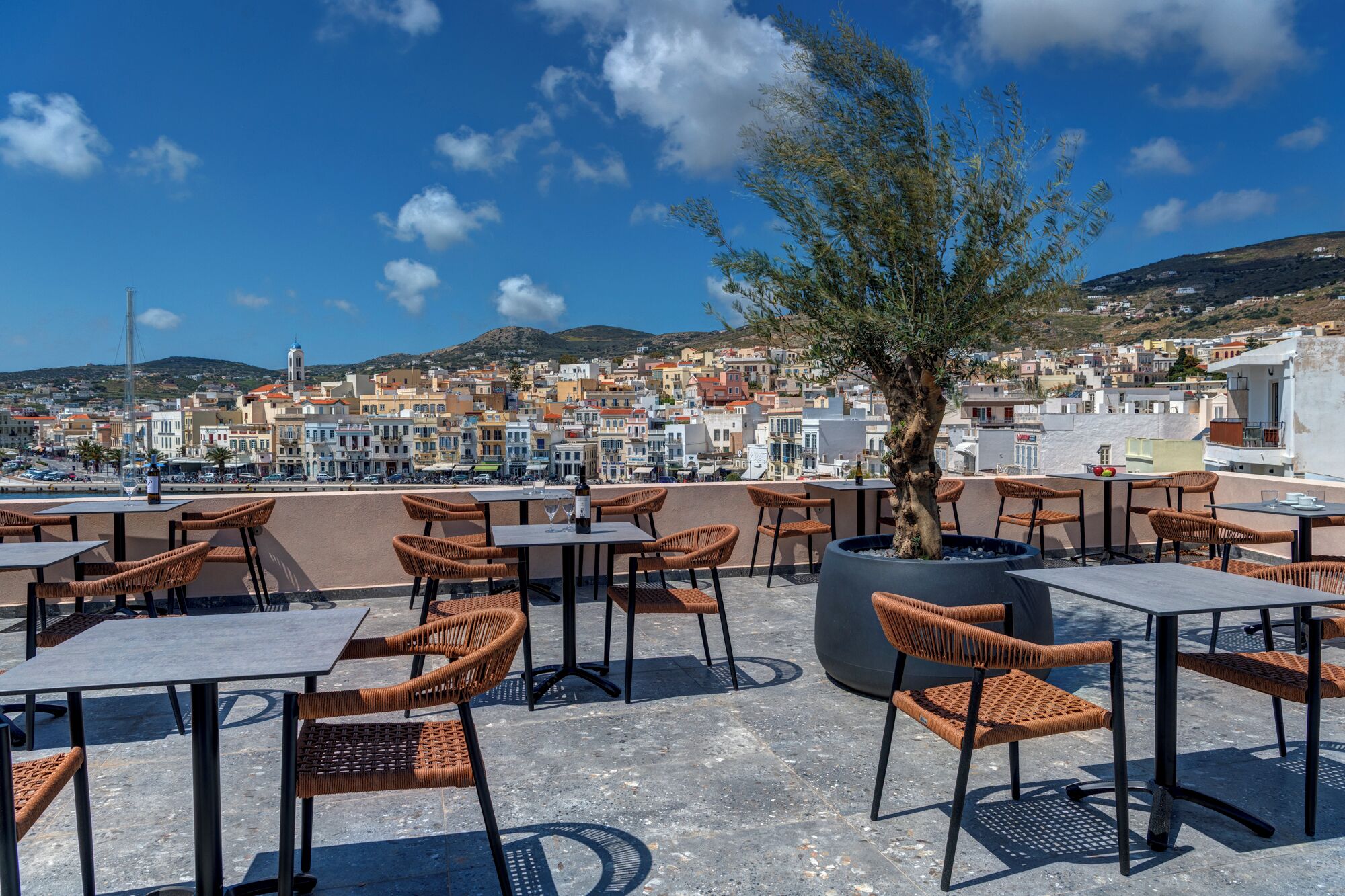 Terraza en la azotea con sillones y mesas de mimbre, con vistas a un pueblo costero con edificios y colinas.