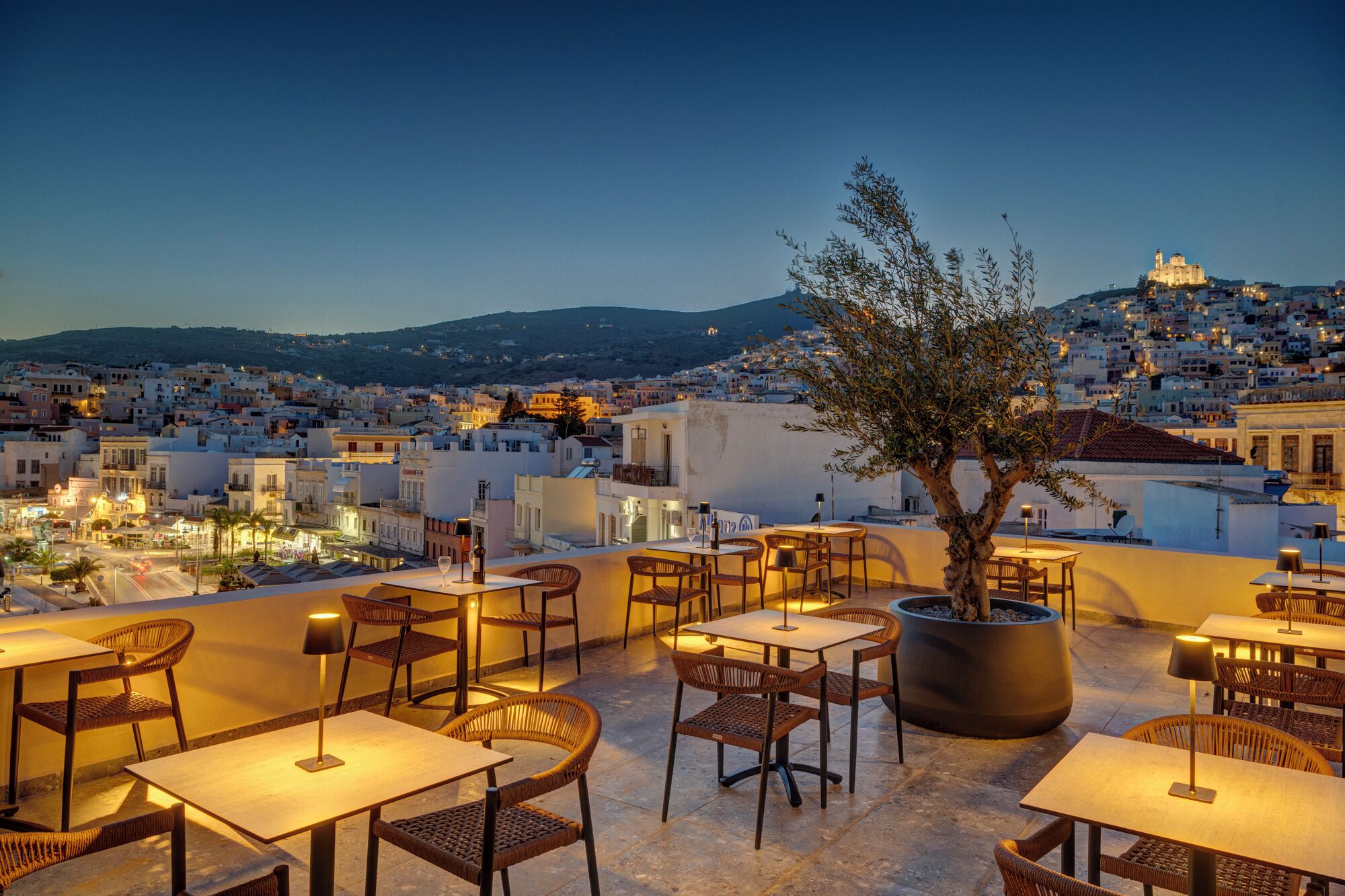 Una terraza en la azotea con mesas y sillas, un gran árbol en maceta y vistas a un paisaje urbano al anochecer.