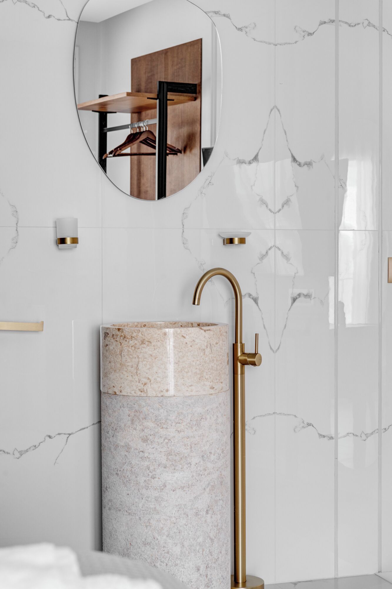 Bany amb un lavabo de marbre, un aixeta daurada i un mirall que reflecteix una prestatgeria de fusta amb roba.