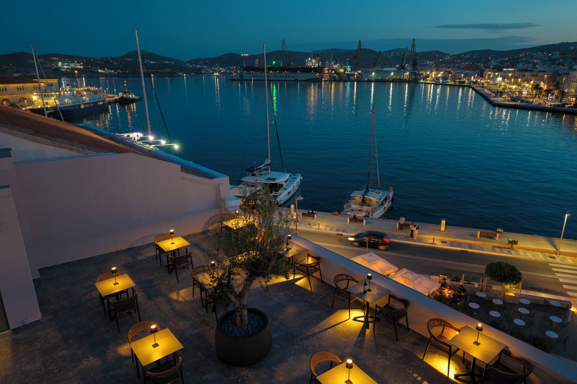 Un espace de restauration en plein air, avec des tables et des chaises, offrant une vue sur un port de plaisance avec des bateaux et sur le paysage urbain en arrière-plan.