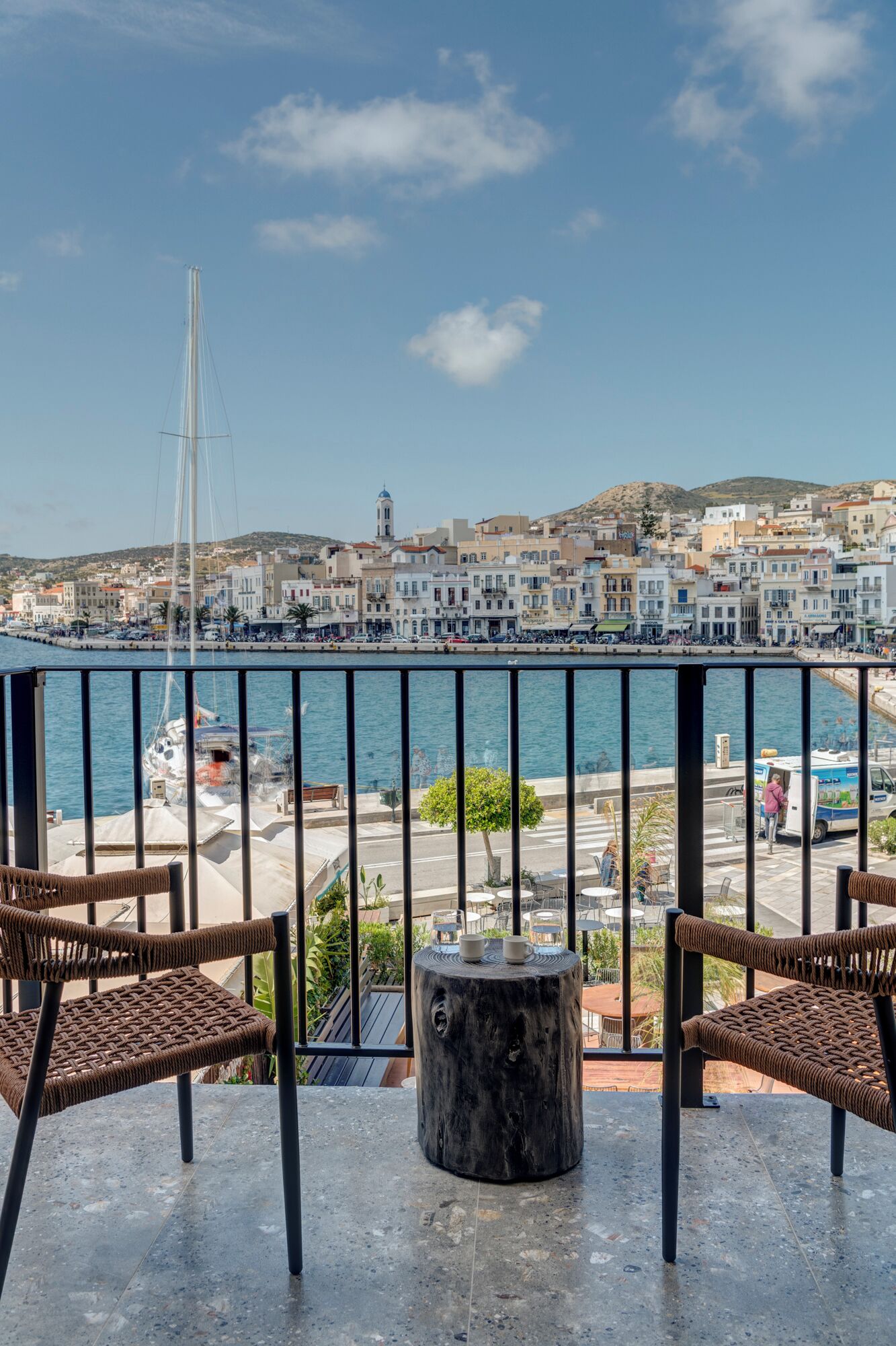 Un balcón con sillas de mimbre y una mesa redonda, con vistas a un puerto con edificios y un velero.