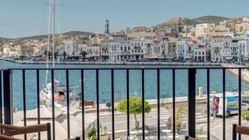 Un balcón con sillas de mimbre y una mesa redonda, con vistas a un puerto con edificios y un velero.