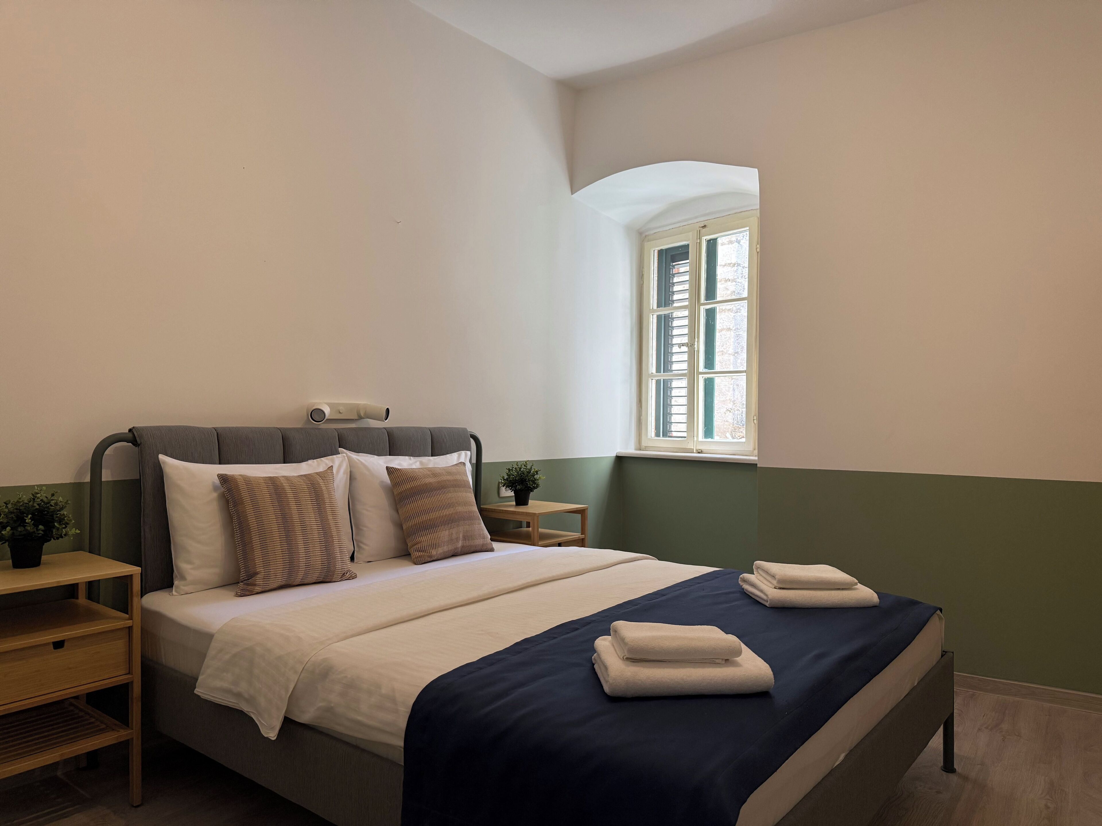 A neatly made bed with a dark blue bedspread, a bedside table with a lamp, and a window with shutters.