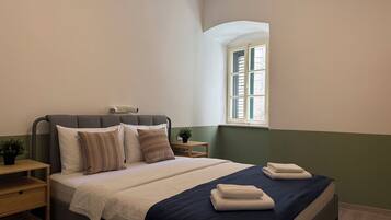 A neatly made bed with a dark blue bedspread, a bedside table with a lamp, and a window with shutters.