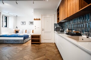 Una habitación de hotel moderna con suelo de madera, una cama con colcha azul, una mesita de noche de madera, una puerta blanca y una zona de cocina con fregadero y estufa.