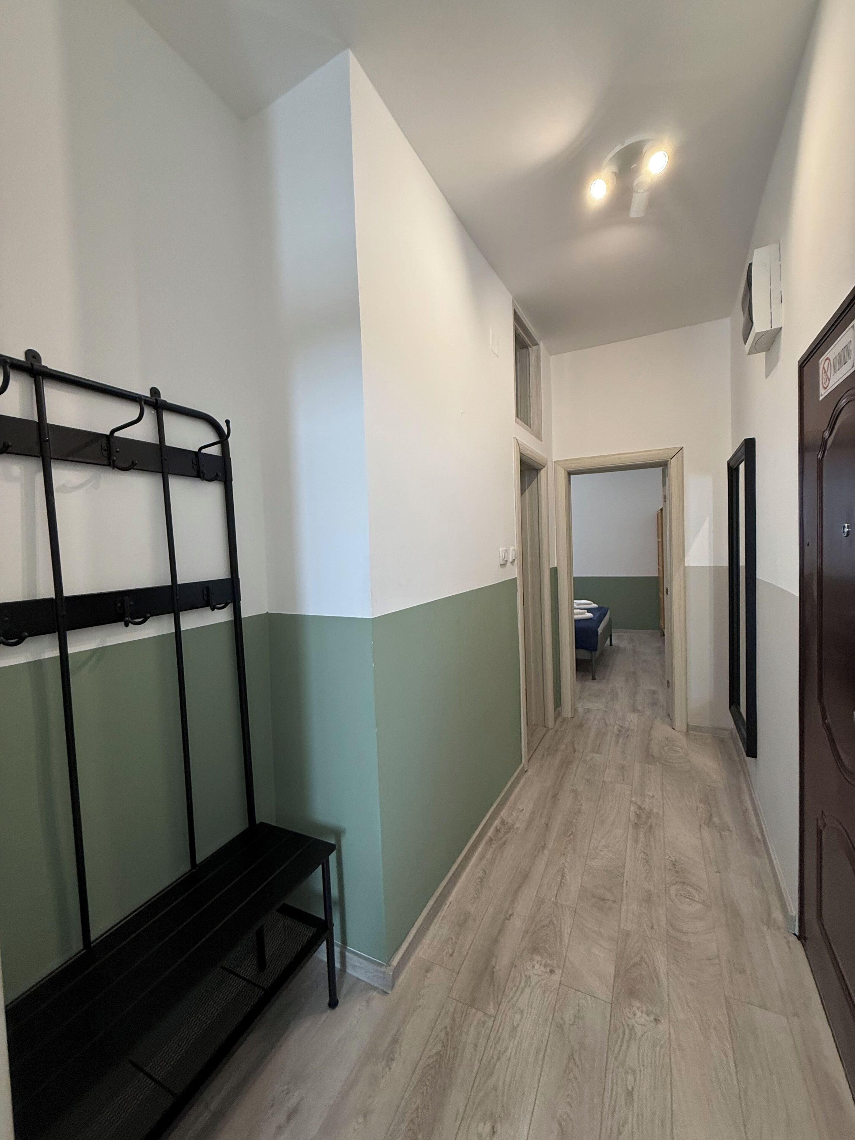 A hallway with a metal bench, a door with a sign, and a bed in the adjacent room.