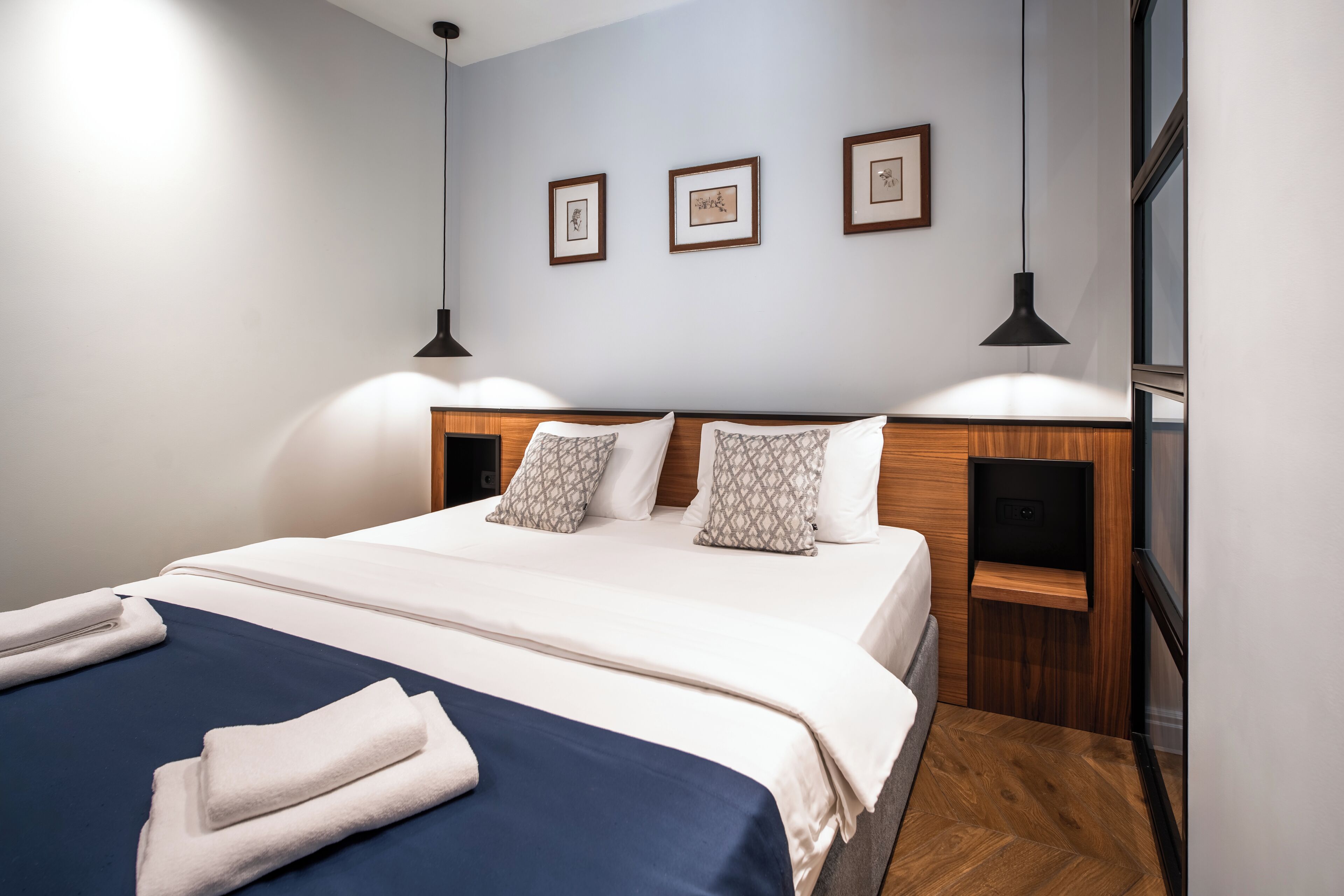 Una cama bien hecha con colcha azul y blanca, dos almohadas y una toalla doblada a un lado. Encima de la cama, hay tres cuadros en la pared.