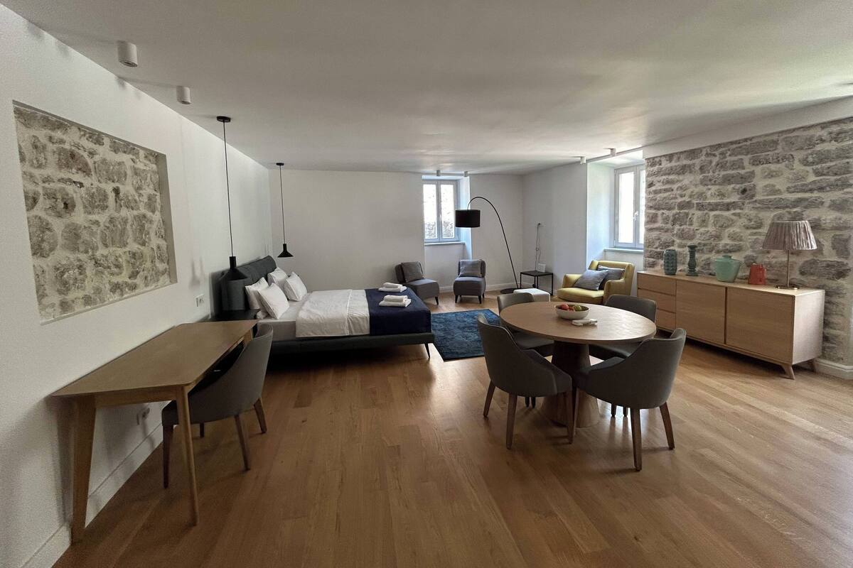 A modern, well-lit room with a bed, a dining table with chairs, a sofa, and a stone wall.