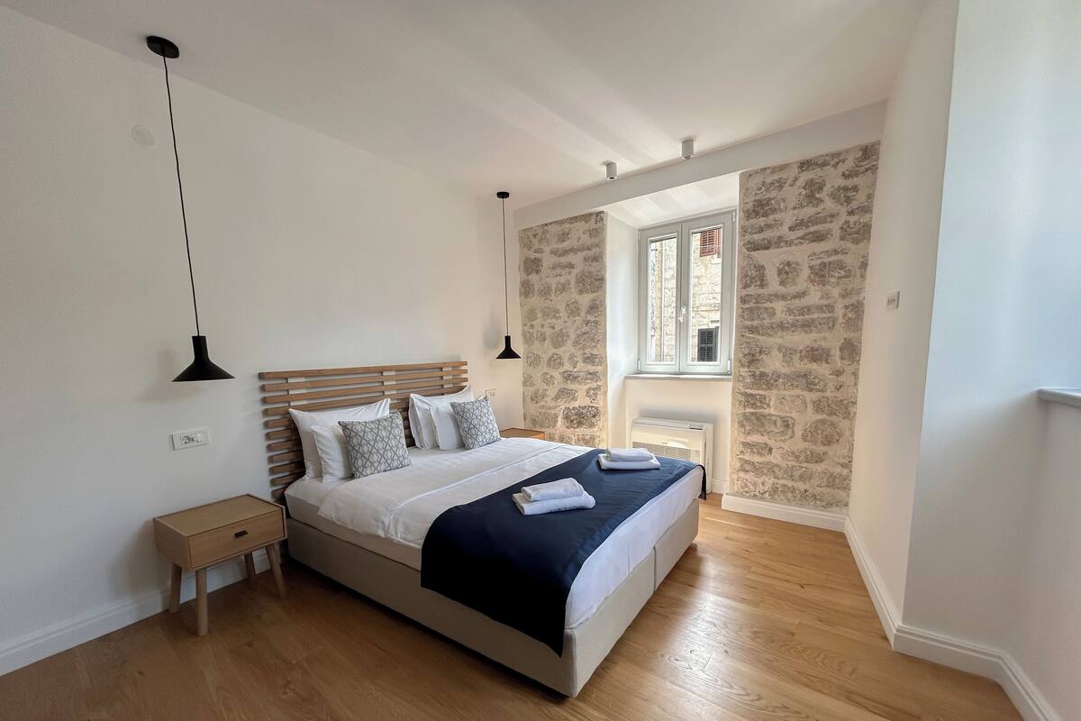 A bedroom with a wooden bed, a nightstand, a stone wall, and a window with a view.