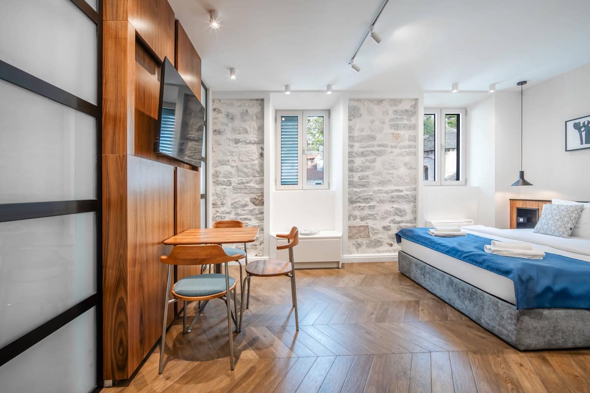 A modern bedroom with a bed, a wooden dining table with chairs, a stone wall, and a window with shutters.