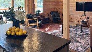 A spacious cabin interior with a wooden ceiling, large windows, a dining area with a table and chairs, and a living room with a sofa and a television.