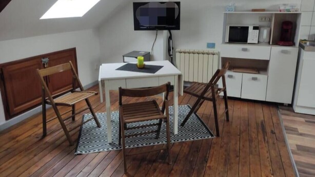 A compact kitchen with a white countertop, a microwave, a kettle, and a small refrigerator. There is a wooden dining table with four chairs and a black and white patterned rug. A television is mounted on the wall above the table. - Le Faubourg (Saint-Quentin)