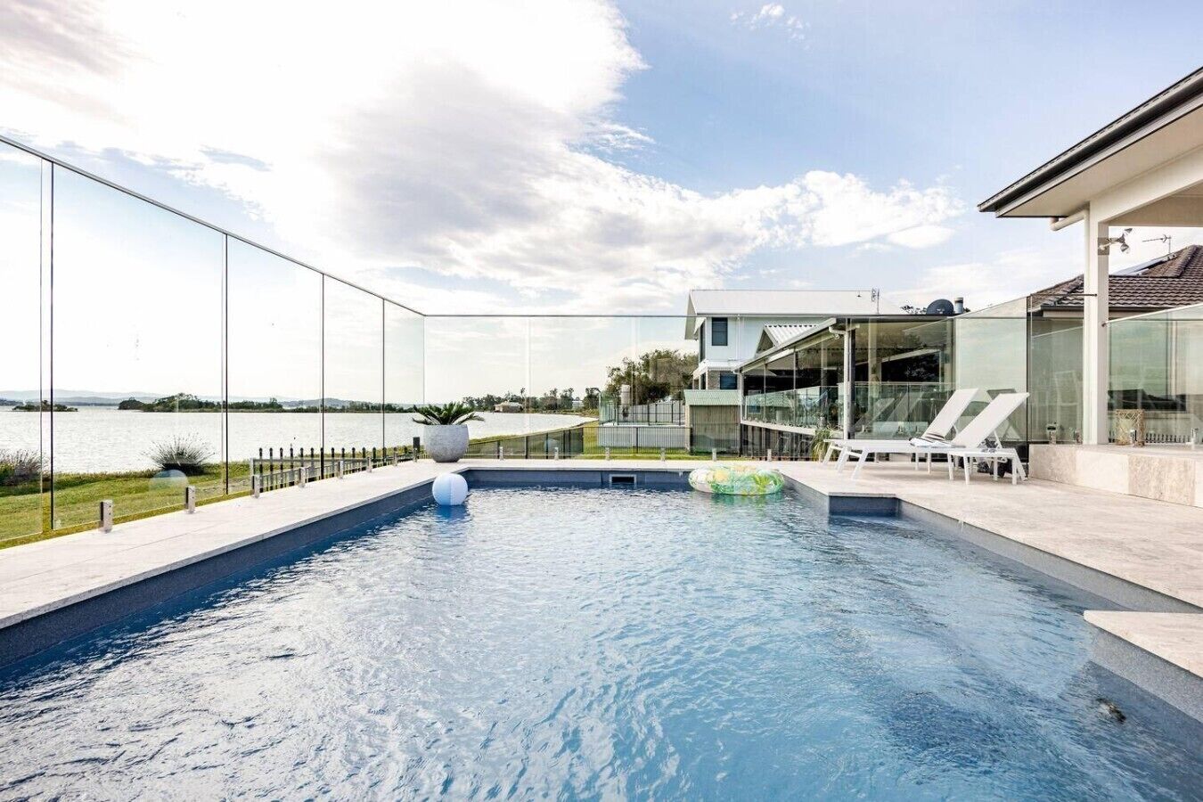 A modern swimming pool with clear glass walls, a view of the water, and a house in the background.