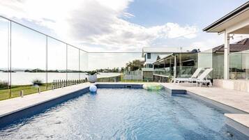 A modern swimming pool with clear glass walls, a view of the water, and a house in the background.