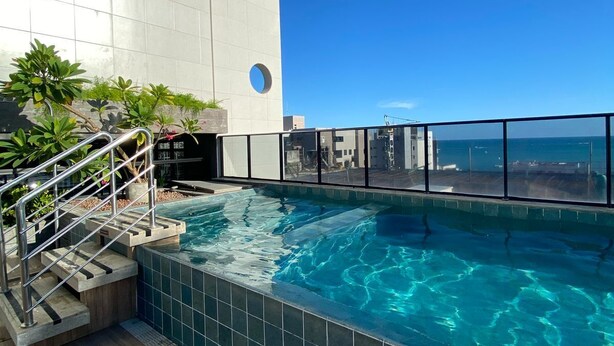 Una piscina en la azotea con agua cristalina y azul, rodeada por un edificio moderno con grandes ventanales y suelo de baldosas.