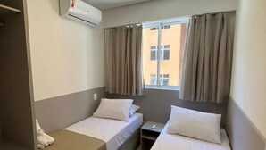 Habitación de hotel con dos camas individuales, un escritorio, una silla, un equipo de aire acondicionado y una ventana con cortinas.