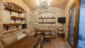 Una habitación acogedora con paredes de piedra, una mesa de madera, sillas y un sofá. Se observan estantes con botellas y un televisor.