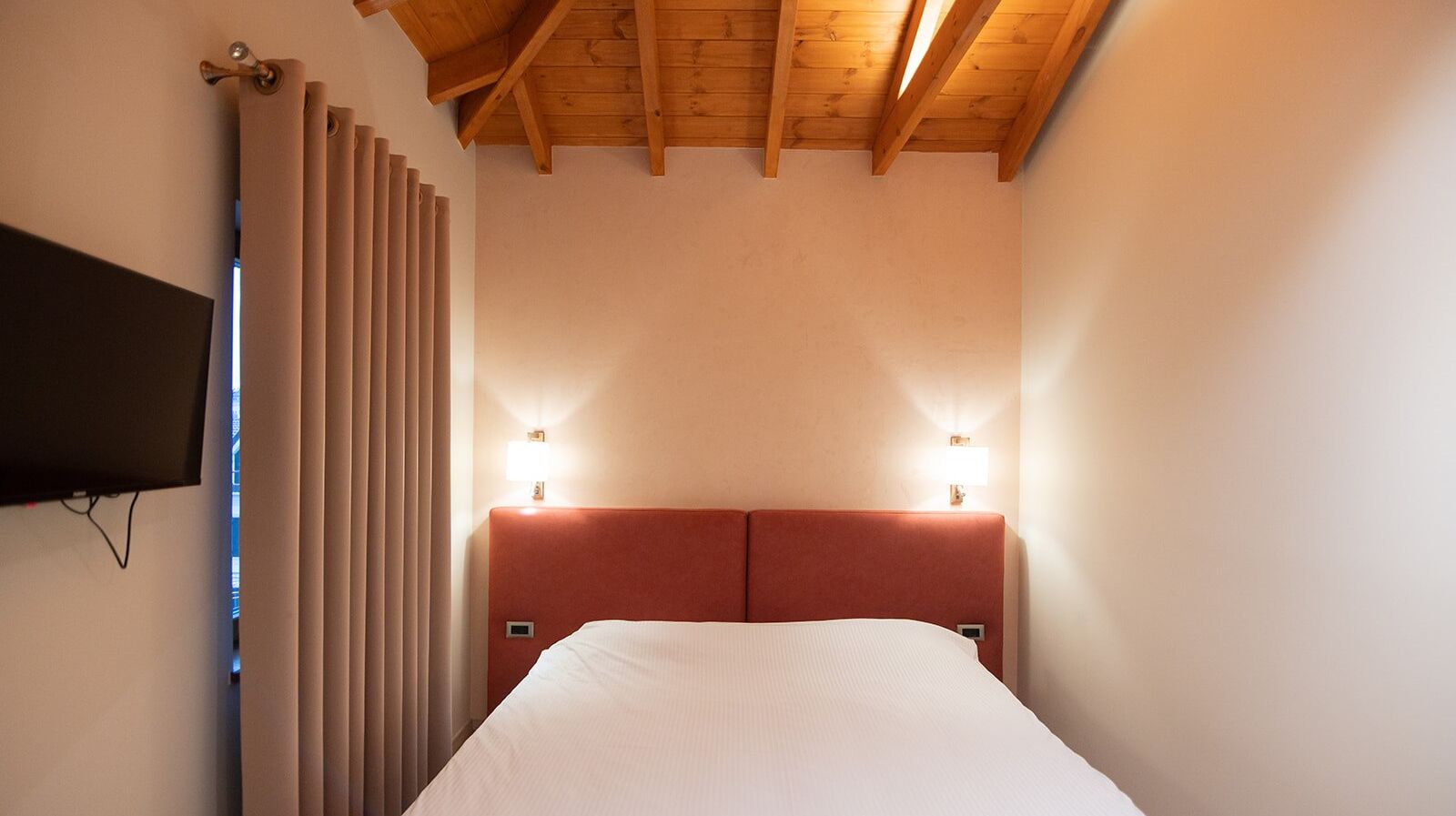 Ett hotellrum med ett träpanelat tak, en säng med en röd sänggavel, två sänglampor och en tv som är monterad på väggen.