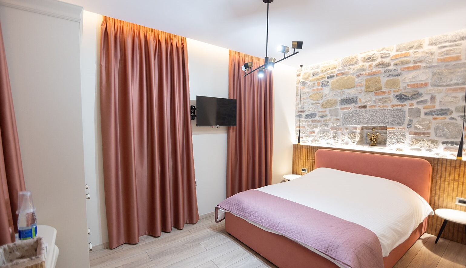A bedroom with a bed, curtains, a television, a stone wall, and a small table.