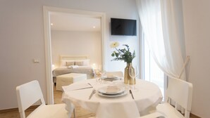 A dining area with a round table set for two, white chairs, a vase with flowers, and a bed in the background. - Ca' Benedetta Suite House Apartsuite Chioggia (Chioggia)