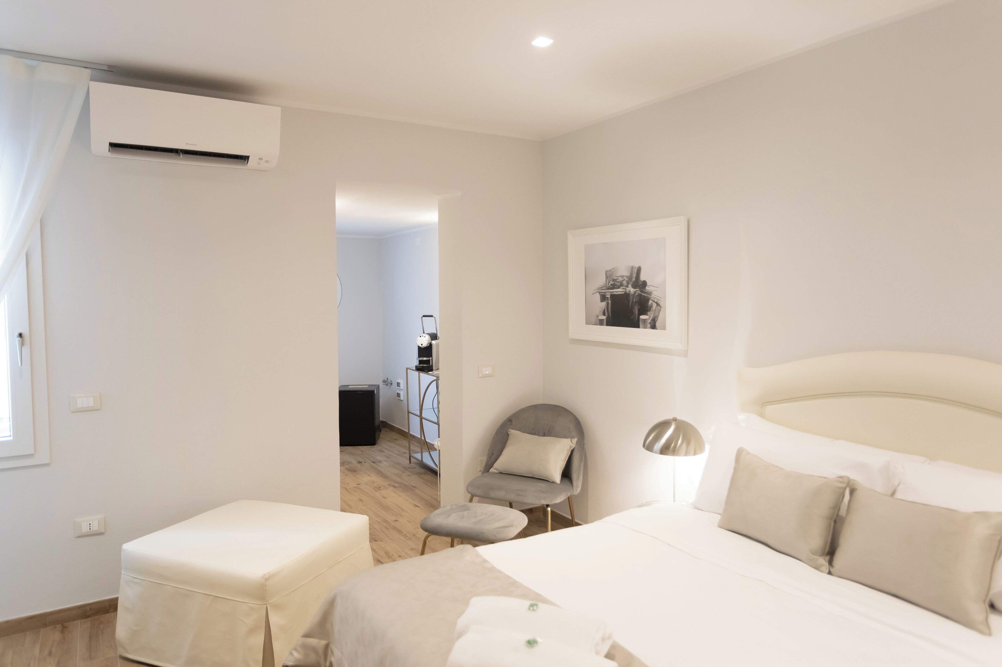 A bedroom with a bed, a chair, an air conditioning unit, a desk, and a framed picture on the wall.