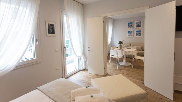 A modern hotel room with a bed, a small table, and a view of the dining area. - Ca' Benedetta Suite House Apartsuite Chioggia (Chioggia)