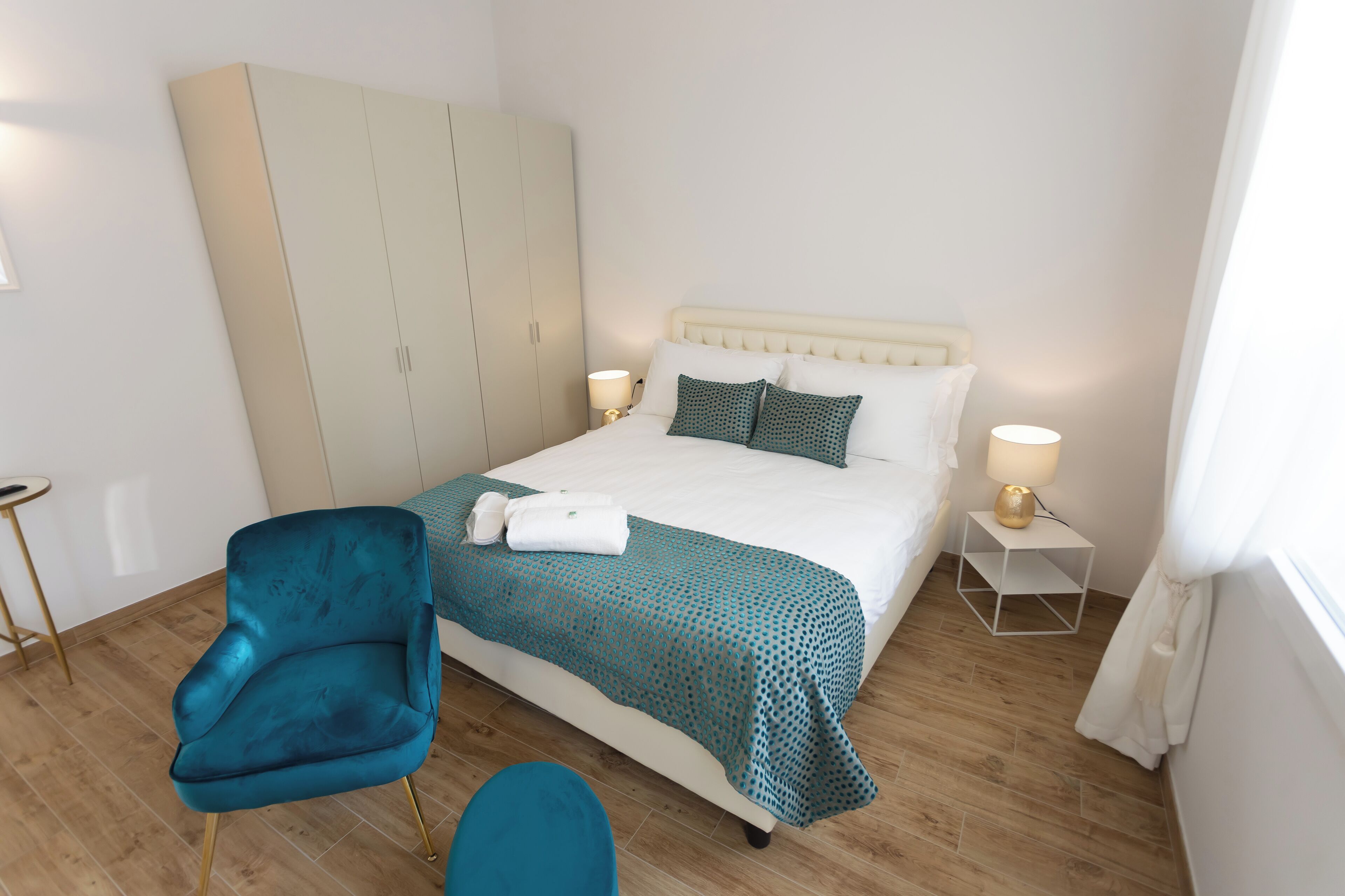 A hotel room with a bed, a wardrobe, a blue chair, a small table, and a nightstand with lamps.