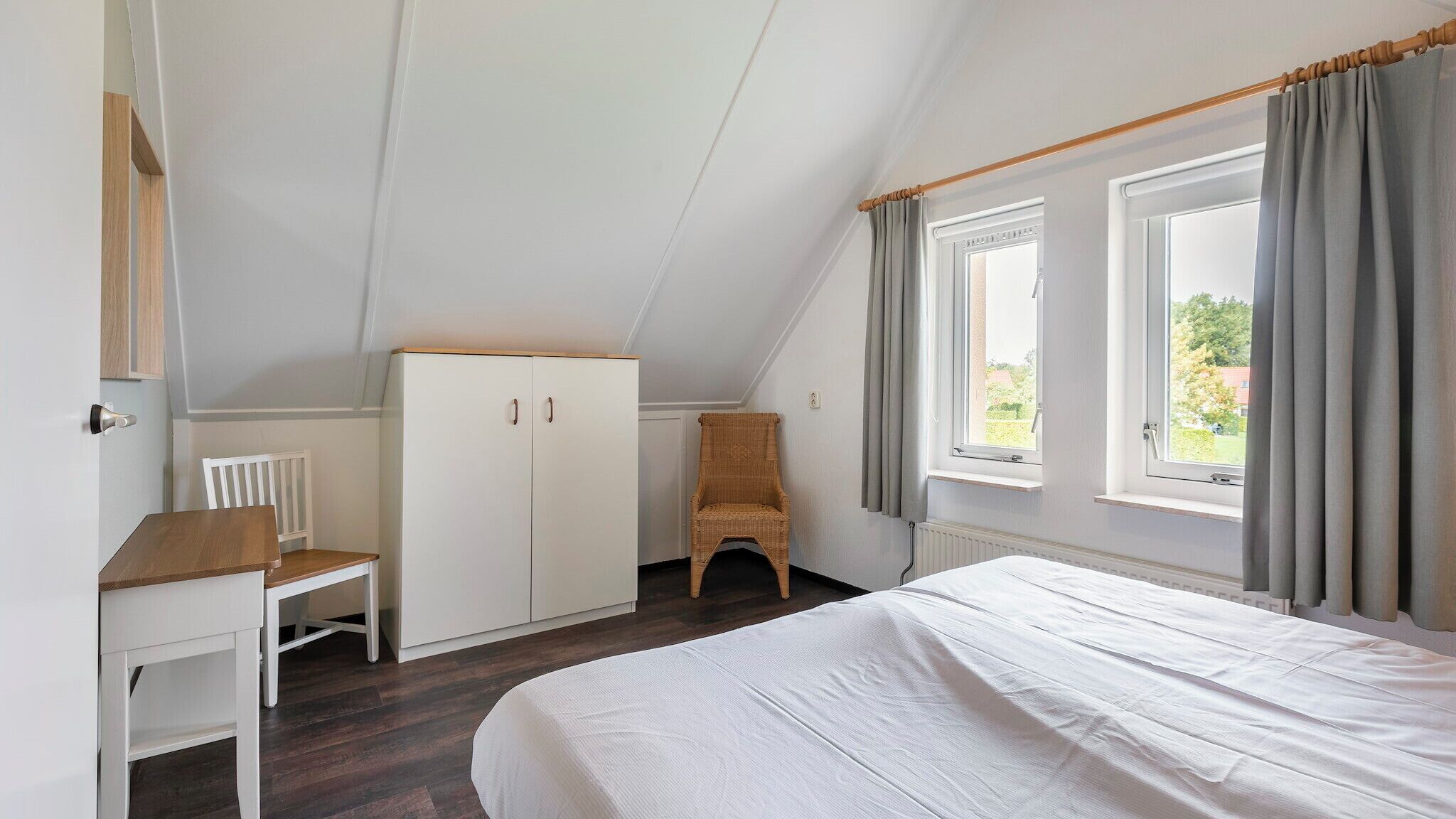 A bedroom with a sloped ceiling, a bed, a wardrobe, a chair, and a window with curtains.