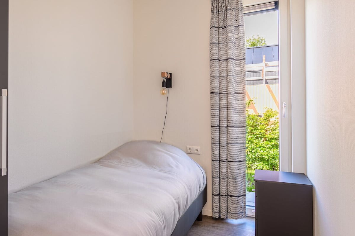 Een kleine, minimalistische slaapkamer met een enkel bed, een stopcontact, een wandlamp en uitzicht op groen buiten.