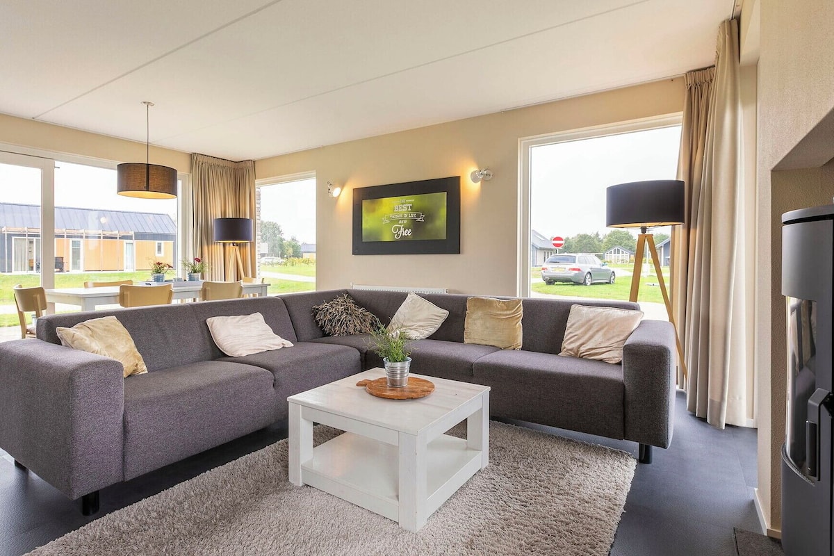 Een moderne woonkamer met een grijze modulaire bank, een witte salontafel en een groot raam met uitzicht op een tuin en gebouwen.