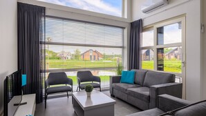 Un salón moderno con un sofá gris, dos sillones, una mesa de centro y un televisor. Amplias ventanas con persianas muestran vista a una zona residencial.