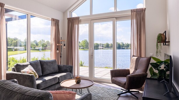 Un salón moderno con un sofá gris, un sillón marrón y un televisor sobre un mueble bajo. Amplias ventanas ofrecen vistas a un lago y vegetación.